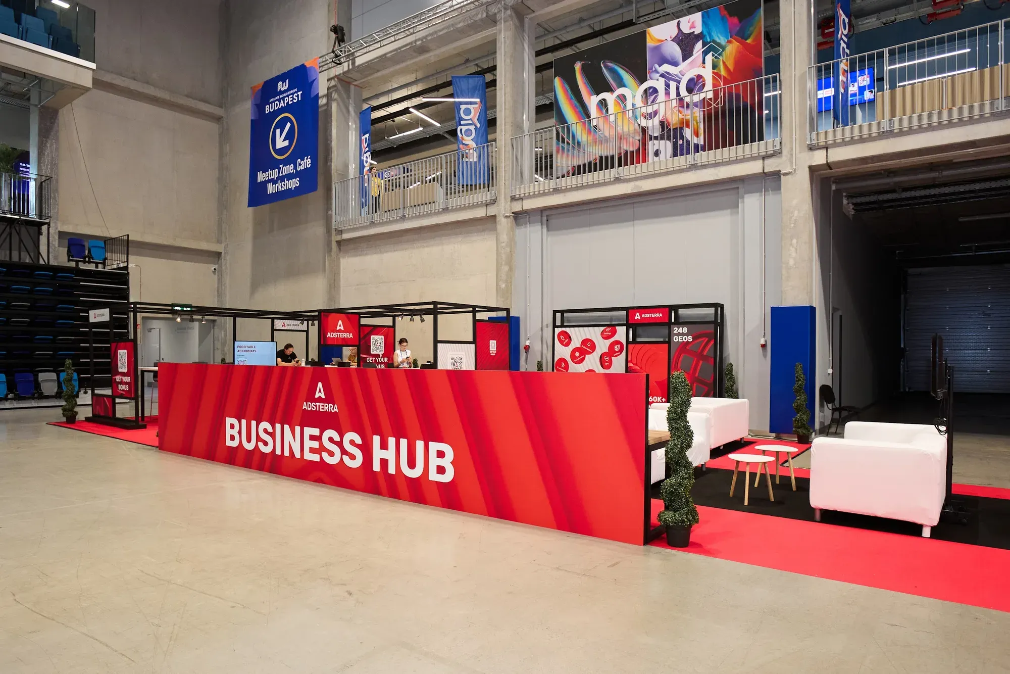 Adsterra Business Hub counter and lounge in the concrete hall at Affiliate World Europe Budapest 2026, under a banner pointing to the Meetup Zone, Café and Workshops