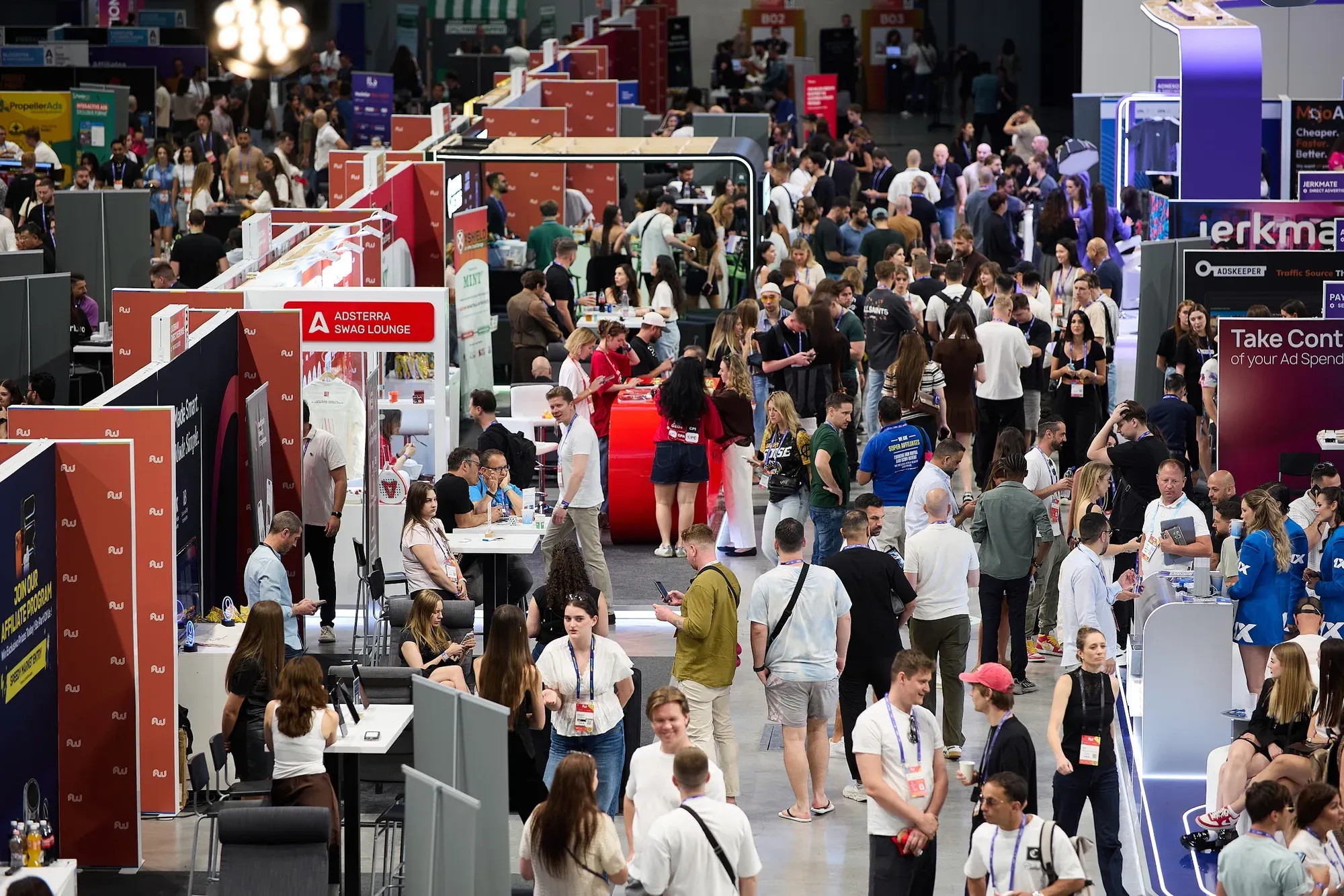 Visitors with lanyards filling the expo floor at Affiliate World Europe 2026 in Budapest, seen from above, with the Adsterra Swag Lounge on the left